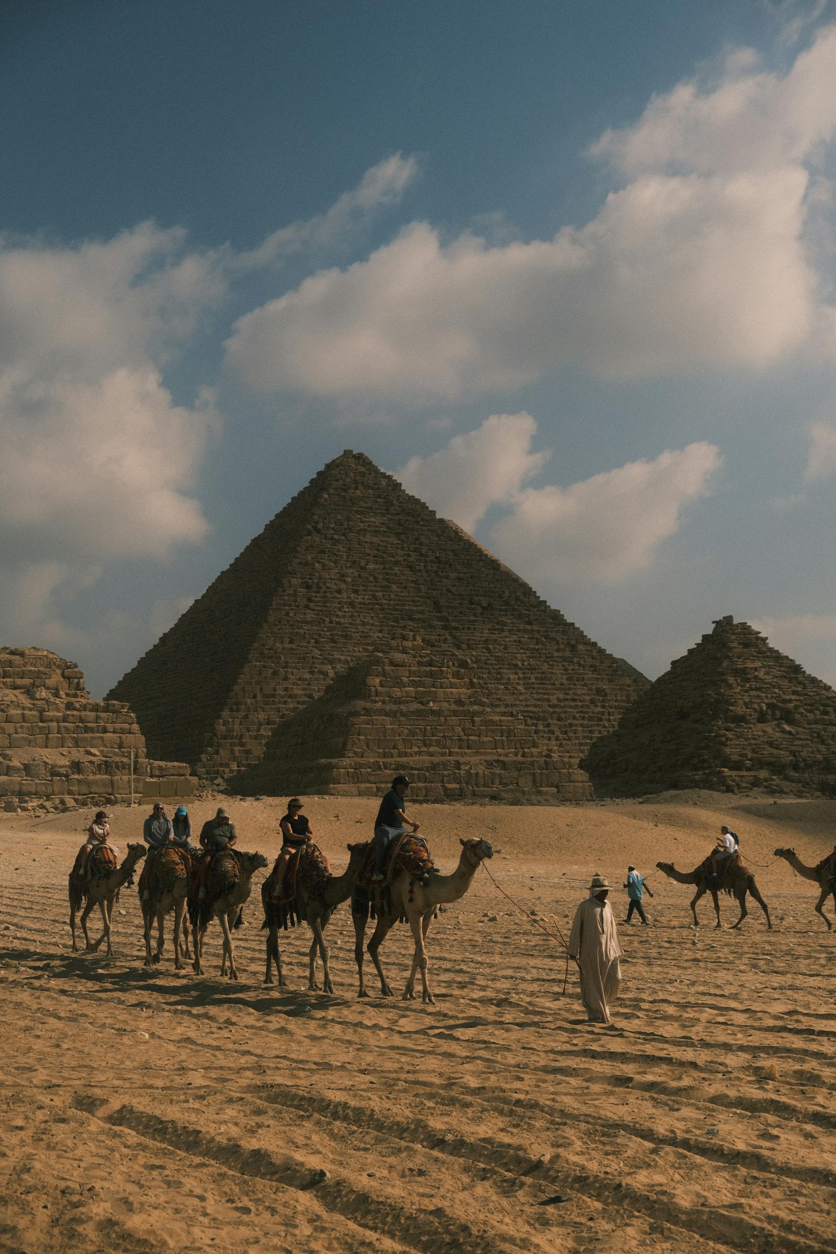 People riding camels in front of the majestic Pyramids of Giza under a cloudy sky.