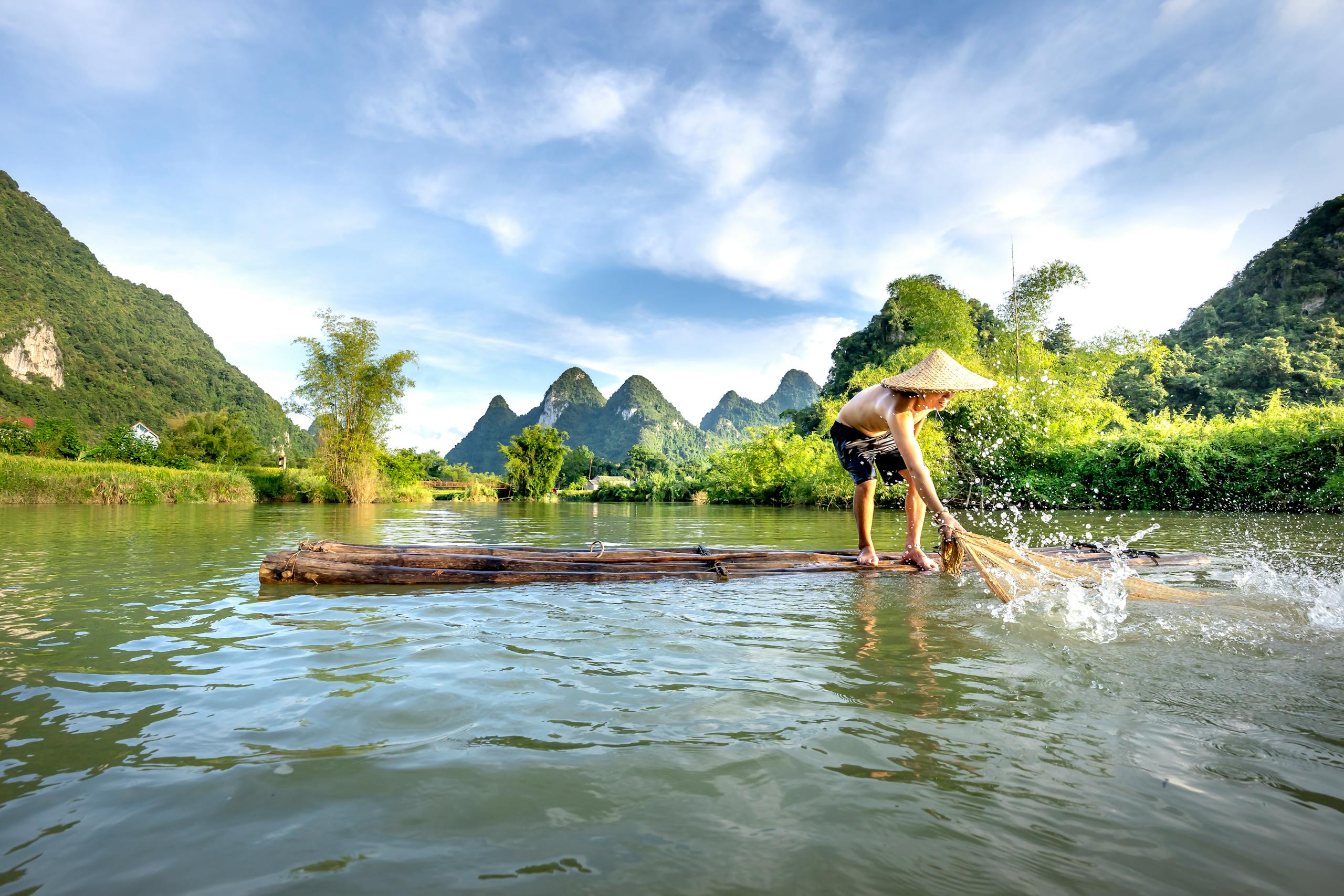 Shirtless man in conical hat paddles a raft on a tranquil river with limestone hills