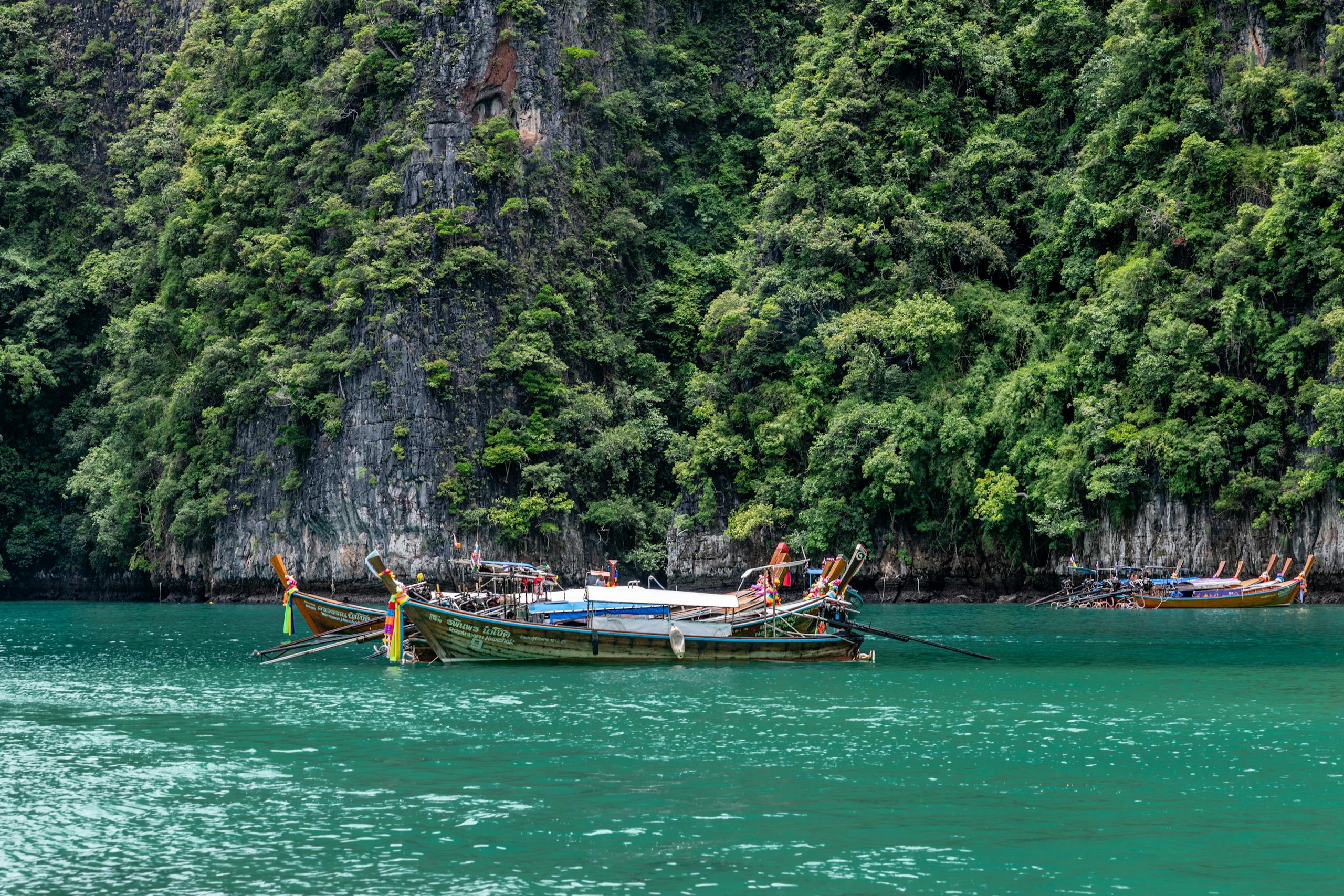 Traditional longtail boats on emerald waters with lush cliffs in Phuket, Thailand, offering a serene tropical view.