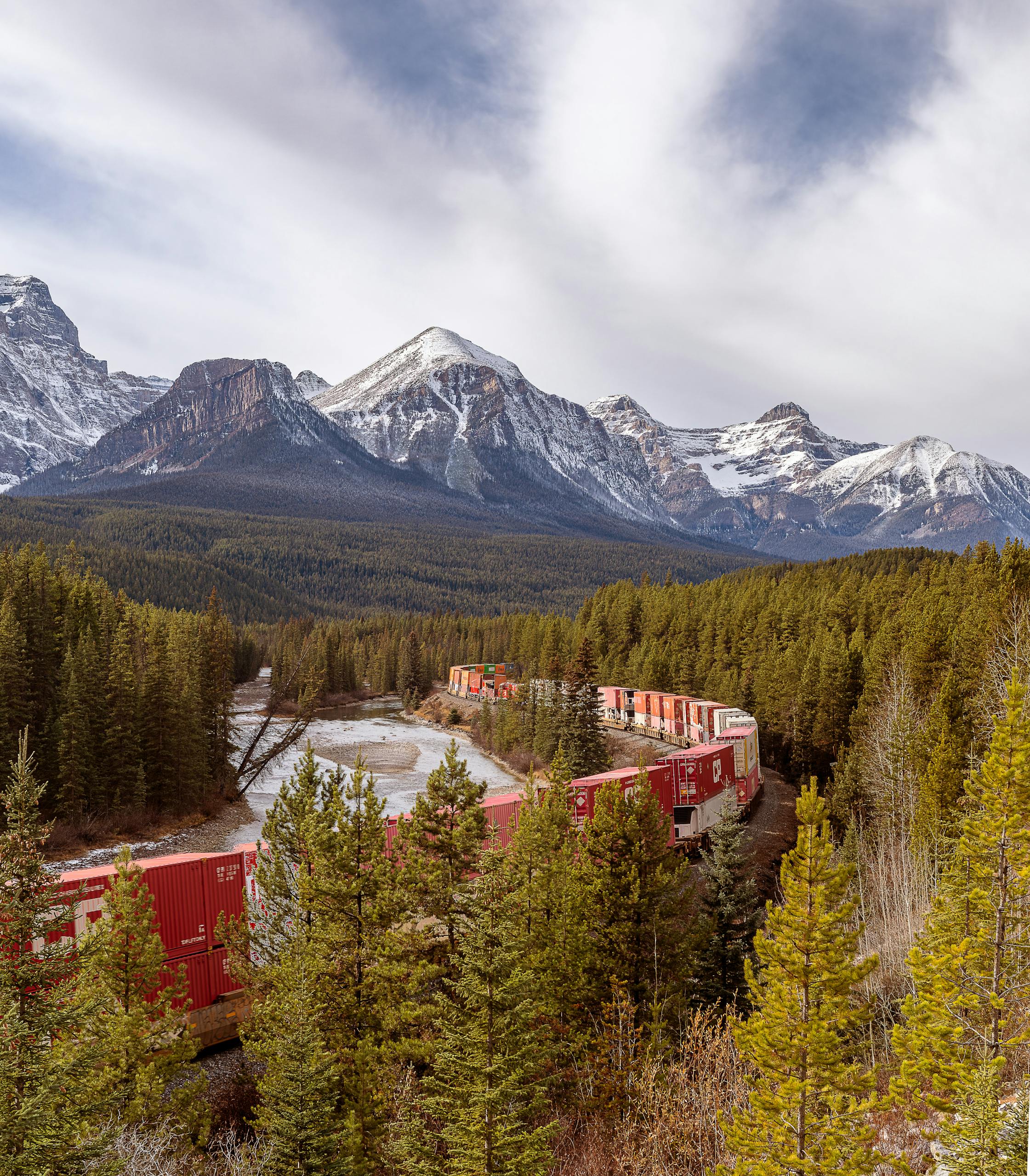 A freight train passing through the scenic Canadian Rockies in Alberta under a dramatic sky.