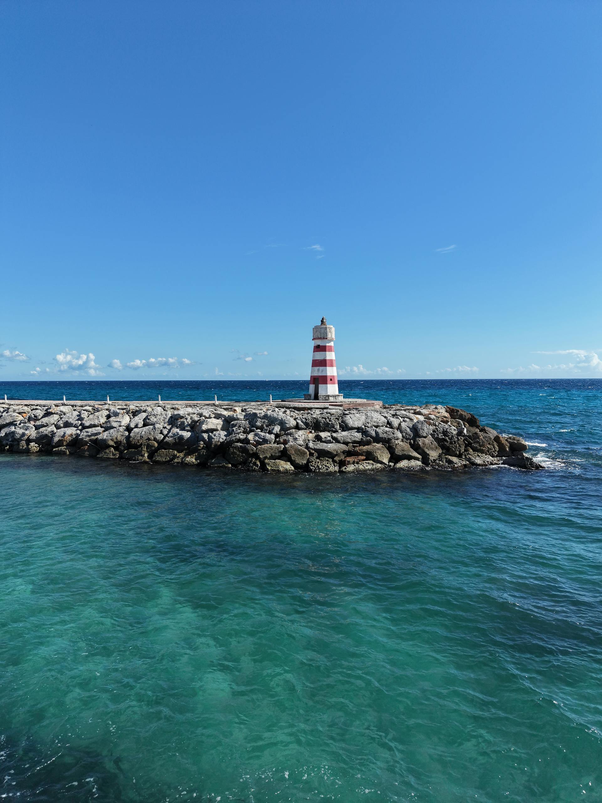 A picturesque striped lighthouse on rocky coast with vibrant Caribbean sea under a clear blue sky.